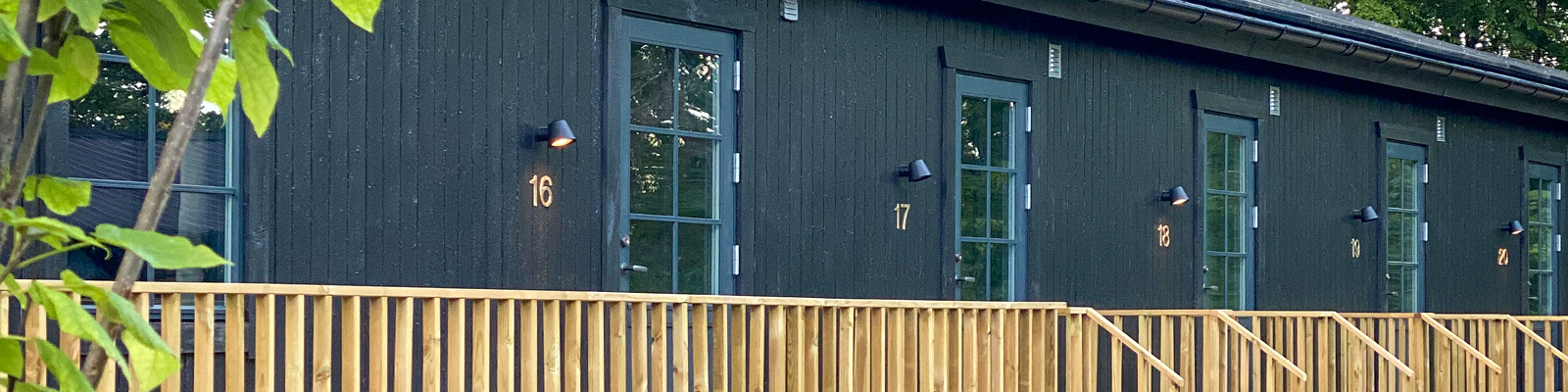 Sort træbygning med numrerede blå døre og træveranda i dagslys.