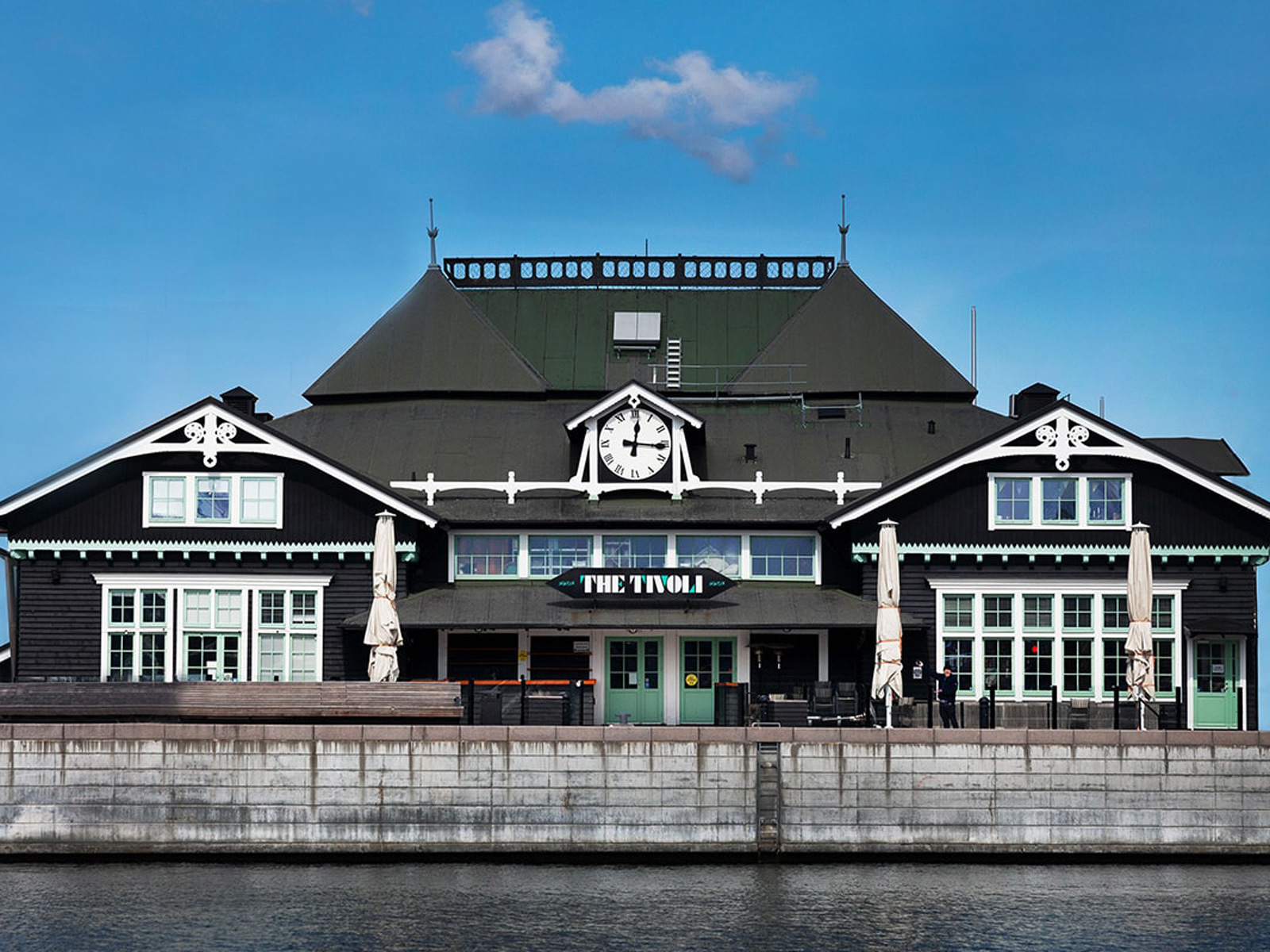 Historisk bygning The Tivoli med sort facade og hvidt ur mod en blå himmel, set forfra ved vandet.