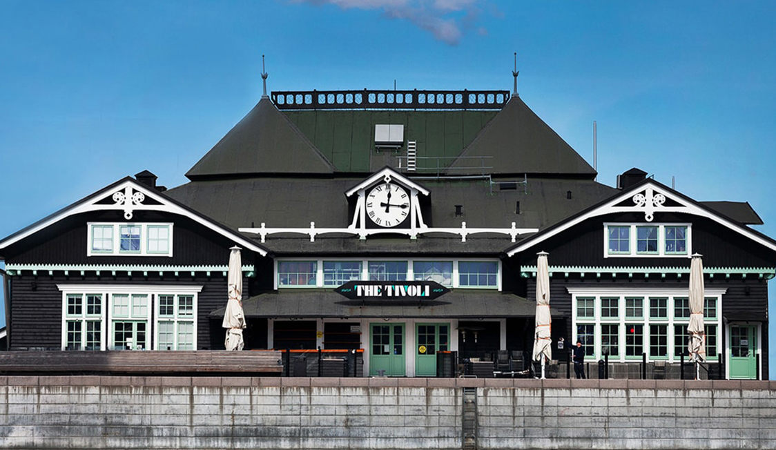 Historisk bygning The Tivoli med sort facade og hvidt ur mod en blå himmel, set forfra ved vandet.