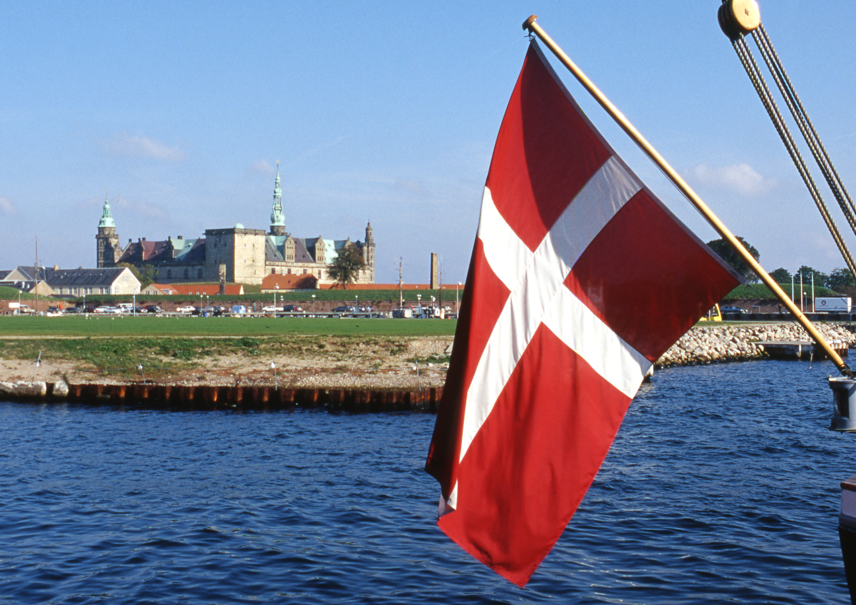 Dansk flag vejer i vinden ved kysten med Kronborg Slot i baggrunden under en klar blå himmel.