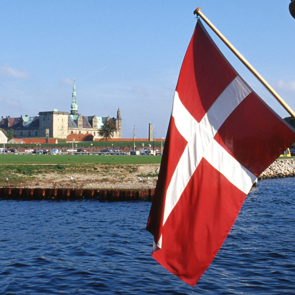 Dansk flag vejer i vinden ved kysten med Kronborg Slot i baggrunden under en klar blå himmel.