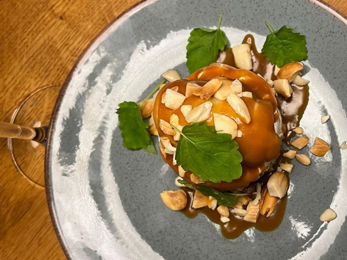 Karamelldessert med hackade nötter och gröna blad på en grå keramiktallrik.