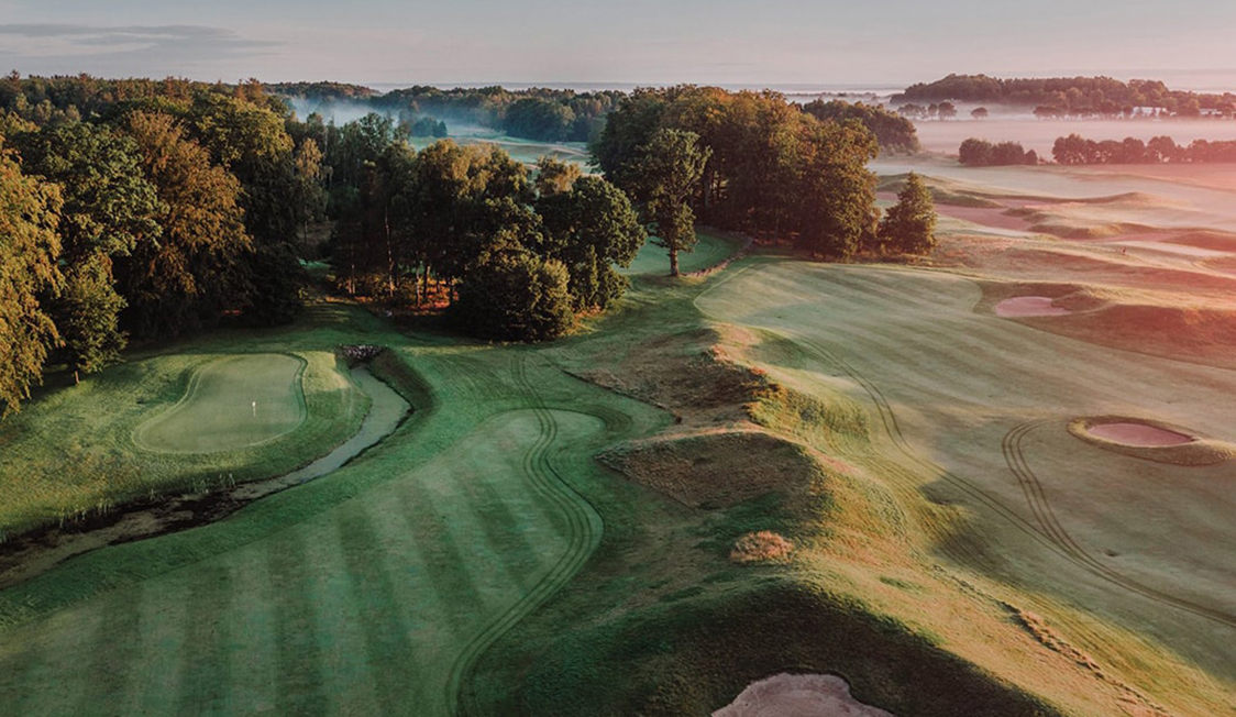 Golfbane set fra oven med frodige grønne fairways og træomkransede greens i morgendis.