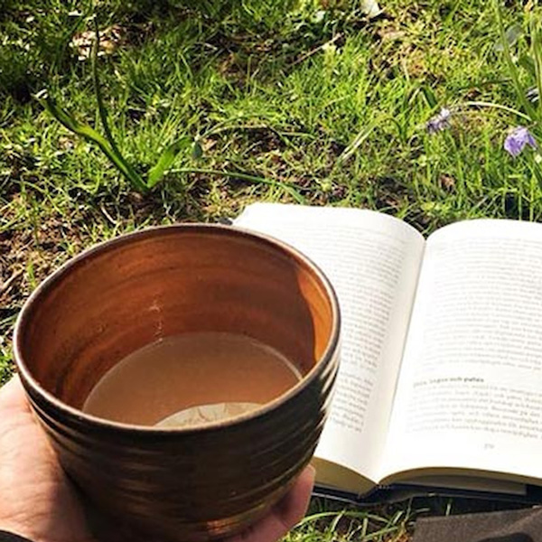 Keramikkop med kaffe holdt i hånd ved siden af en åben bog i græsset.