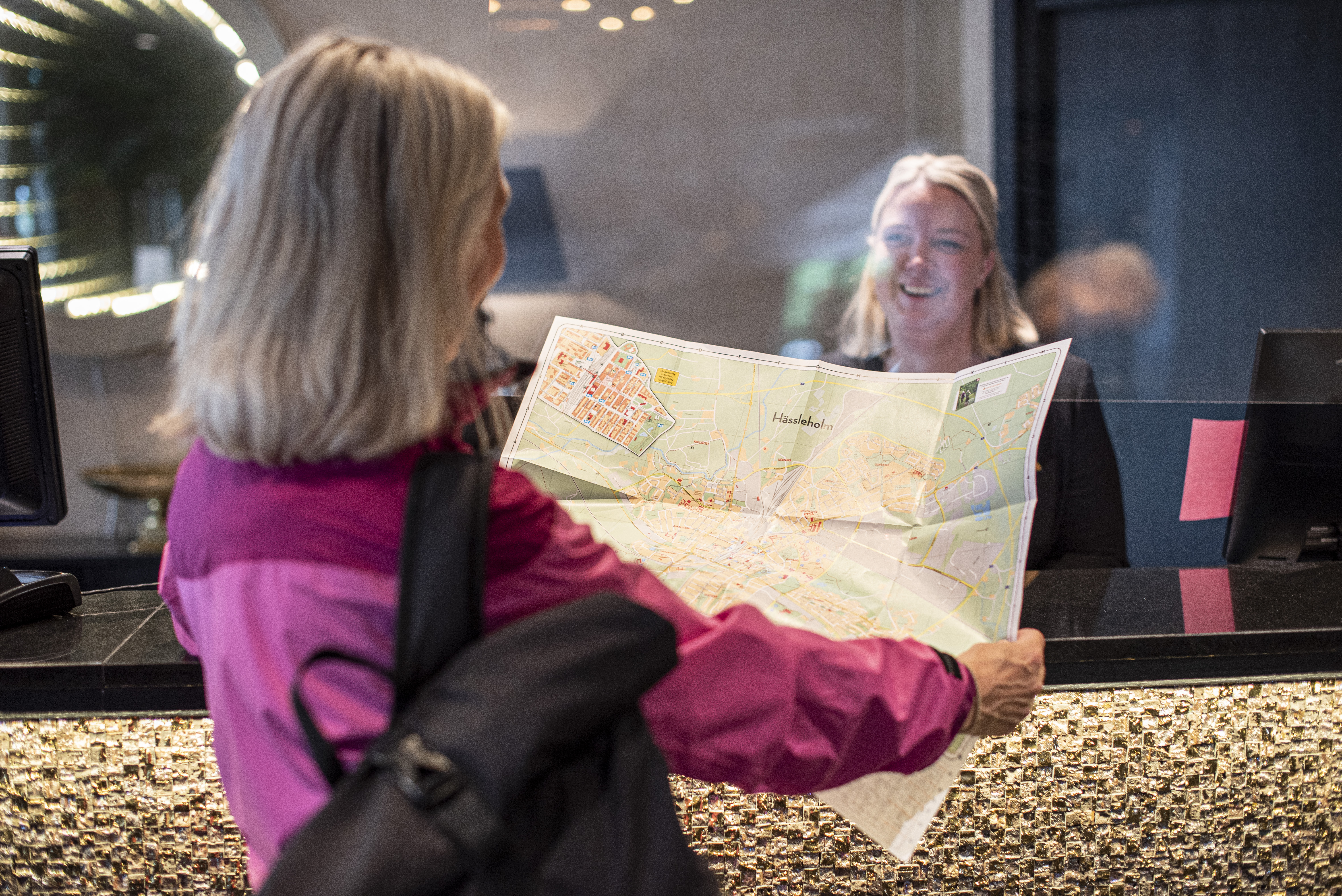 Person i pink jakke viser kort over Hässleholm til receptionist i hotellets lobby.