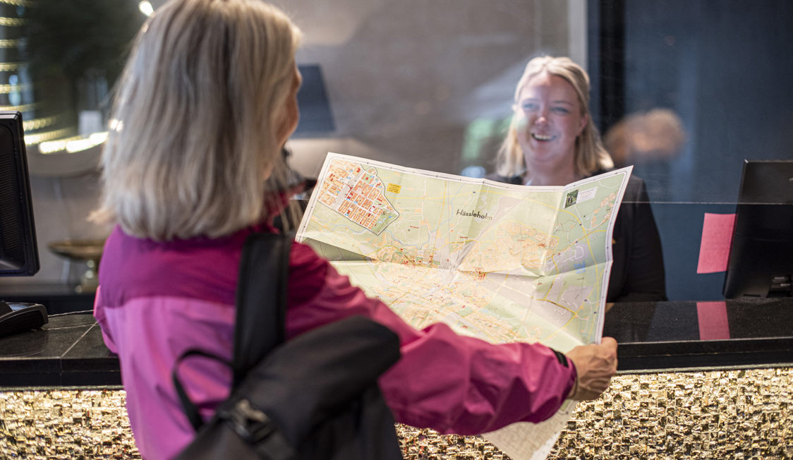 Person i pink jakke viser kort over Hässleholm til receptionist i hotellets lobby.