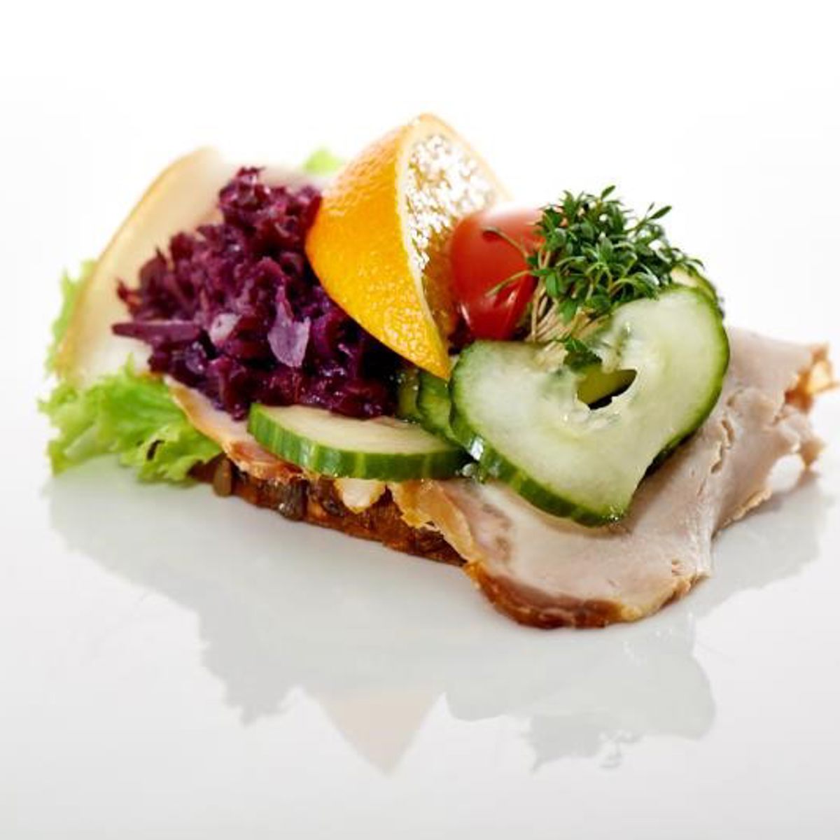 Smørrebrød med flæskesteg, rødkål, skiver af agurk, appelsin, tomat og friske krydderurter på en skive rugbrød.