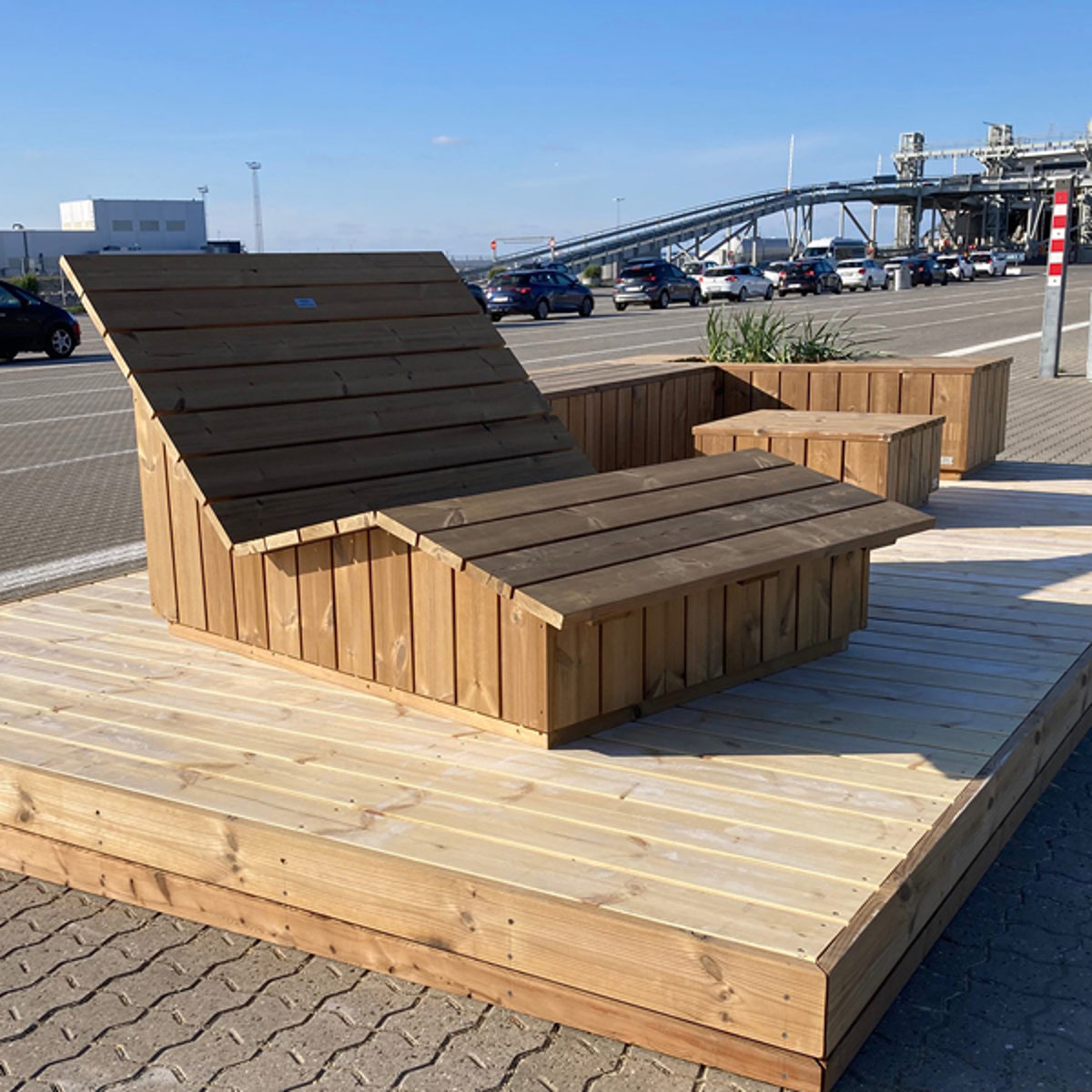 Træbænk med liggefunktion på en platform ved en vej, designet til udendørs afslapning i offentlige områder.