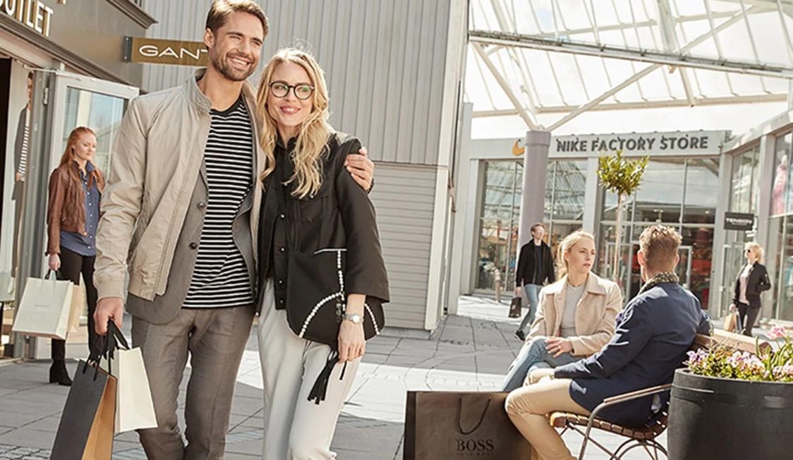 Par smilende mennesker med shoppingposer fra tøjbutikker i et udendørs outlet-center på en solskinsdag.