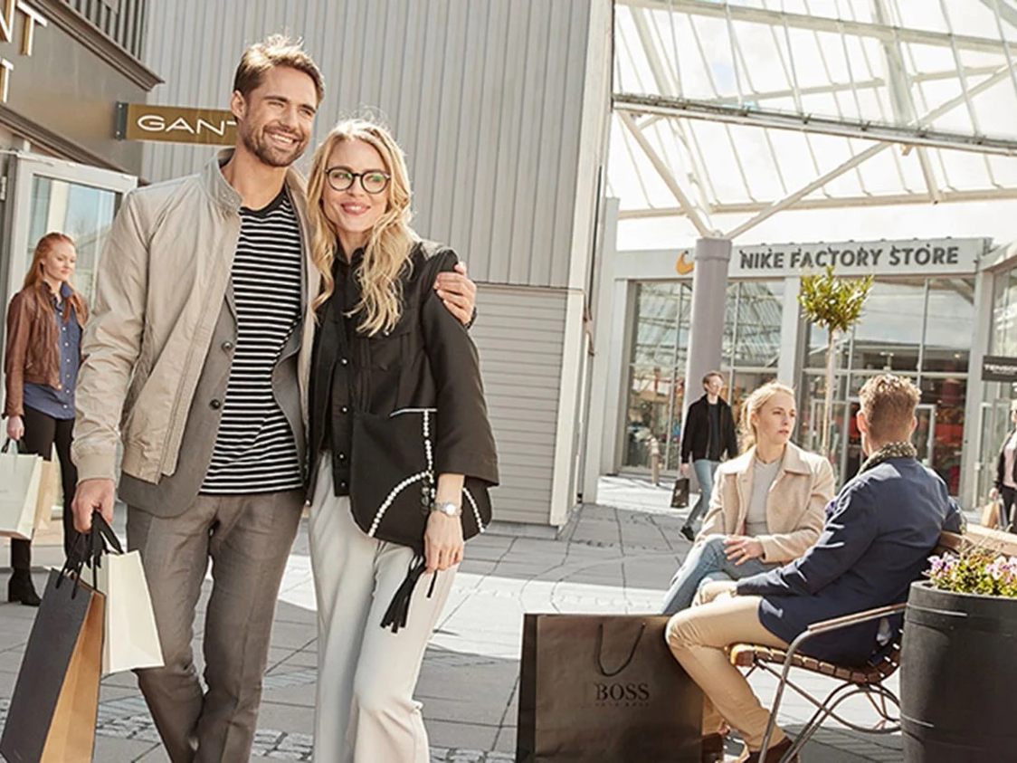 Par smilende mennesker med shoppingposer fra tøjbutikker i et udendørs outlet-center på en solskinsdag.