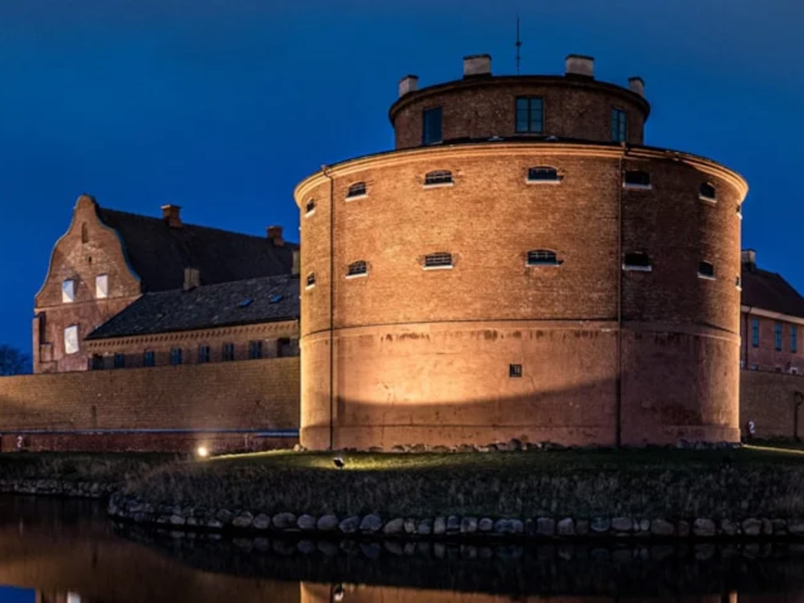 Rundt fæstningstårn af mursten oplyst ved nat langs vandkanal i Sverige.