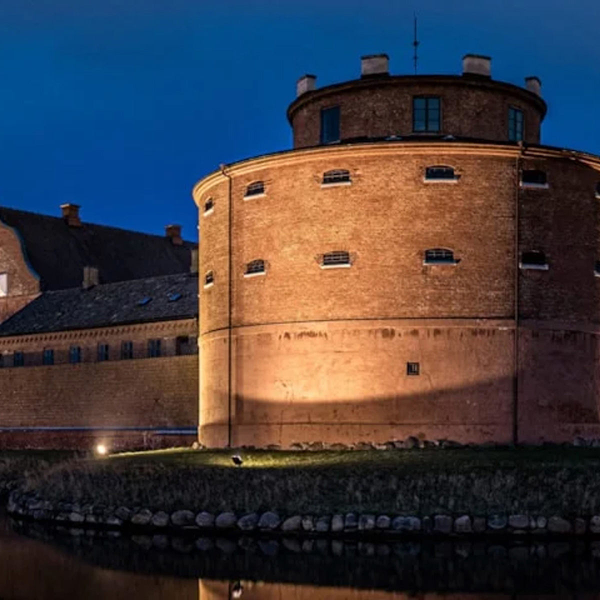 Rundt fæstningstårn af mursten oplyst ved nat langs vandkanal i Sverige.