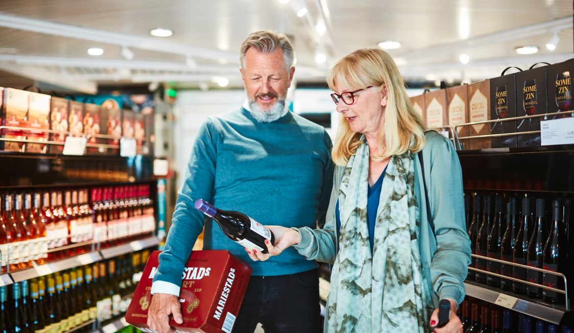Ældre par ser på rødvinsflaske i skibets vinbutik, omringet af flasker på hylder.