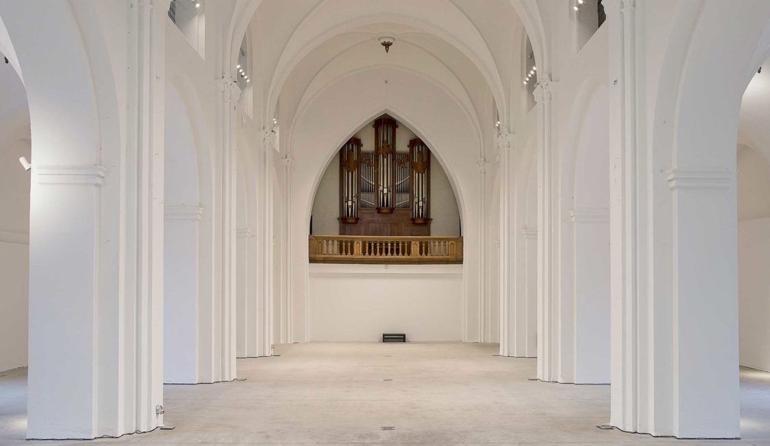 Stort hvidt kirkerum med høje buede søjler og imponerende orgel midt i baggrunden i minimalistisk stil.