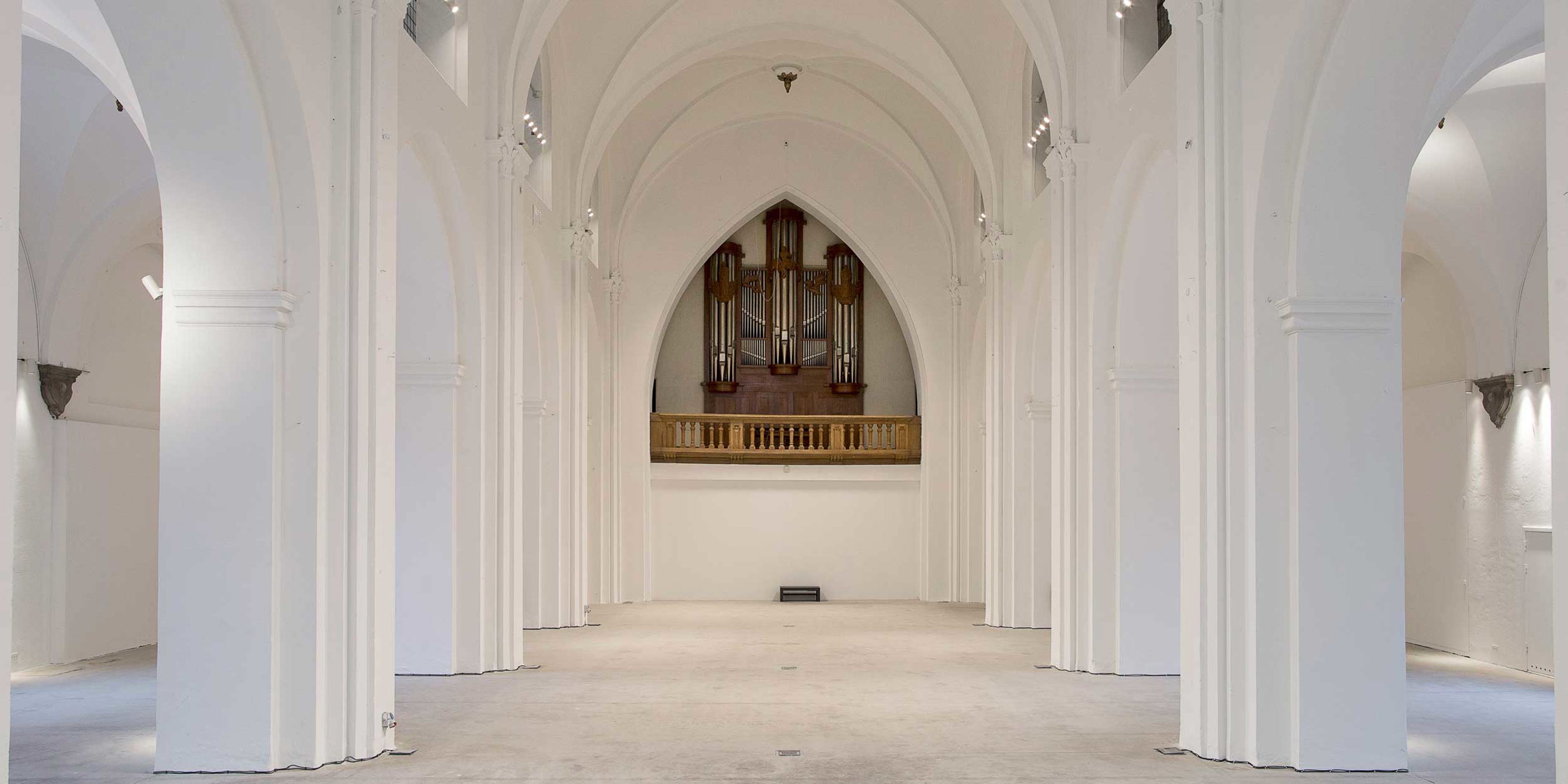 Stort hvidt kirkerum med høje buede søjler og imponerende orgel midt i baggrunden i minimalistisk stil.