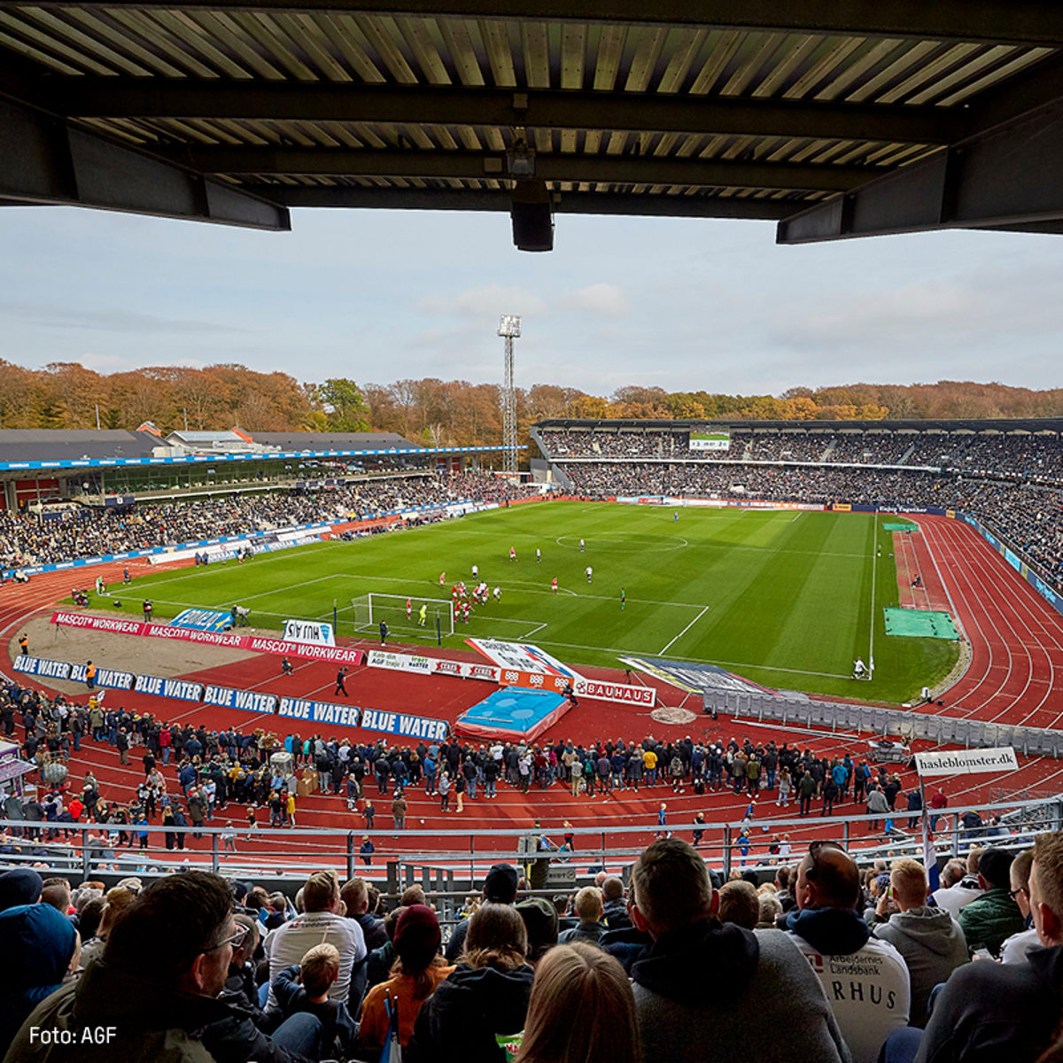Fodboldkamp i gang på Ceres Park stadion set fra tribunen, med fyldt publikum og efterårslandskab i baggrunden.