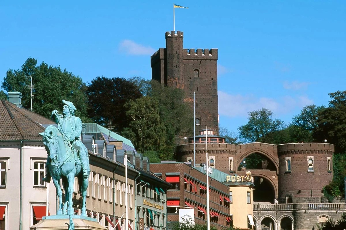 Rytterstatue foran middelalderborg i solskin, med tårne og grønne træer som baggrund.