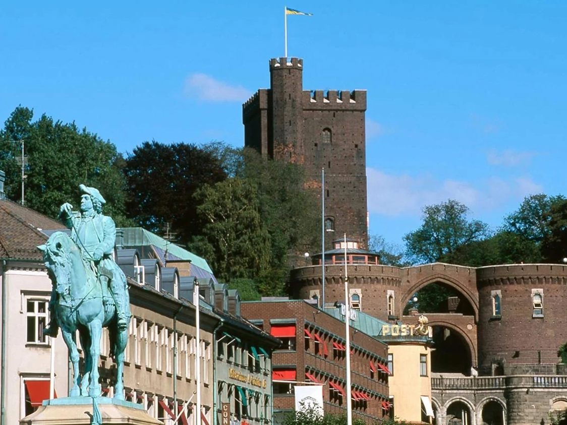 Rytterstatue foran middelalderborg i solskin, med tårne og grønne træer som baggrund.