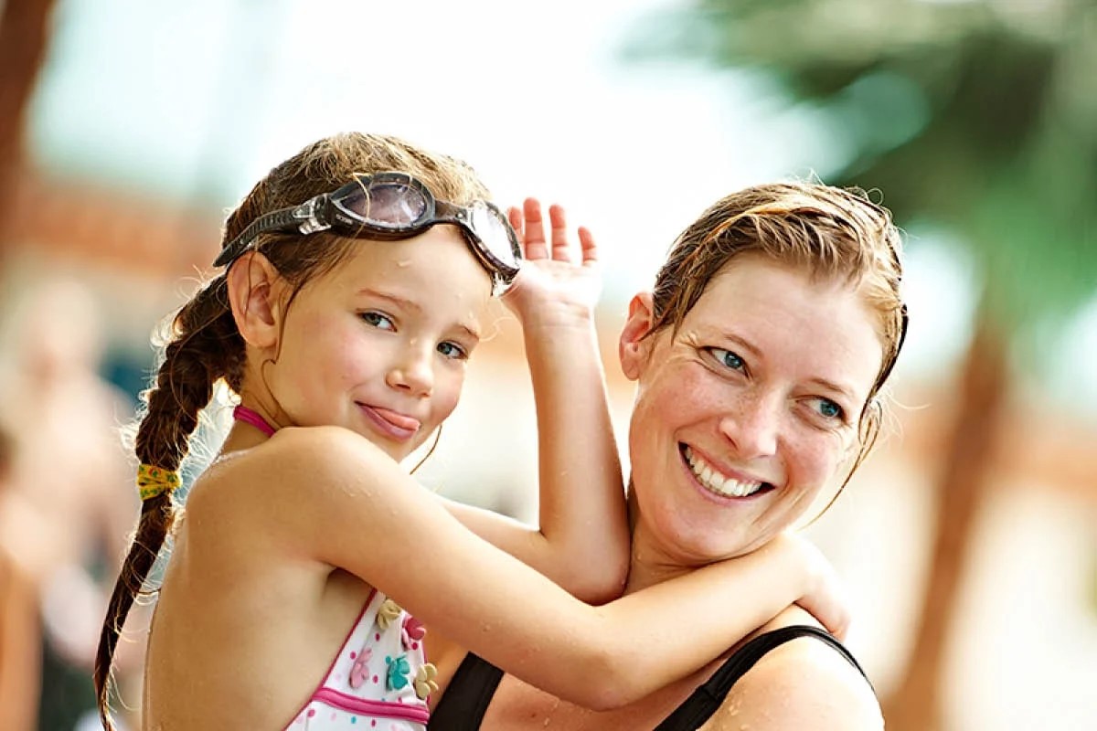 Mor och dotter skrattar tillsammans på vattenpark, dottern bär simglasögon på huvudet.