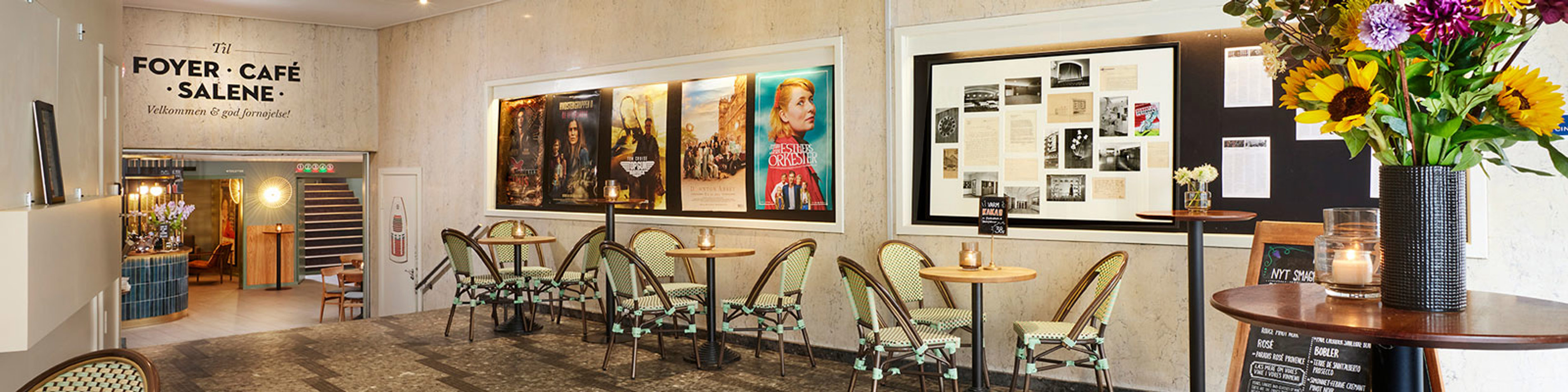 Café i en foyer med elegante træborde, blomstervaser og filmplakater på væggen, set i et hyggeligt miljø.