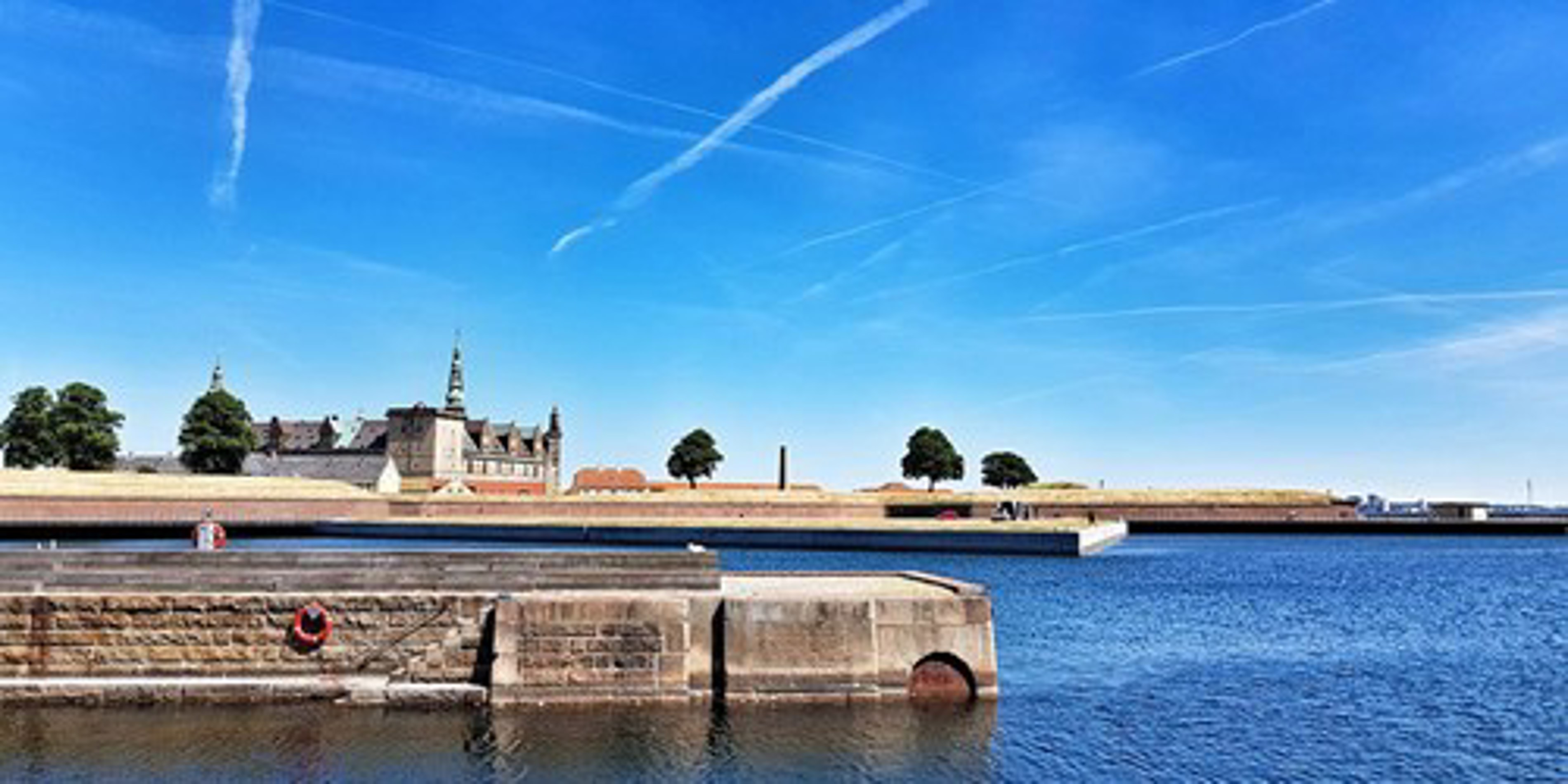 Kronborg Slot ved kysten i Helsingør, set fra vandet med blå himmel og kontrasterende vandspejl.