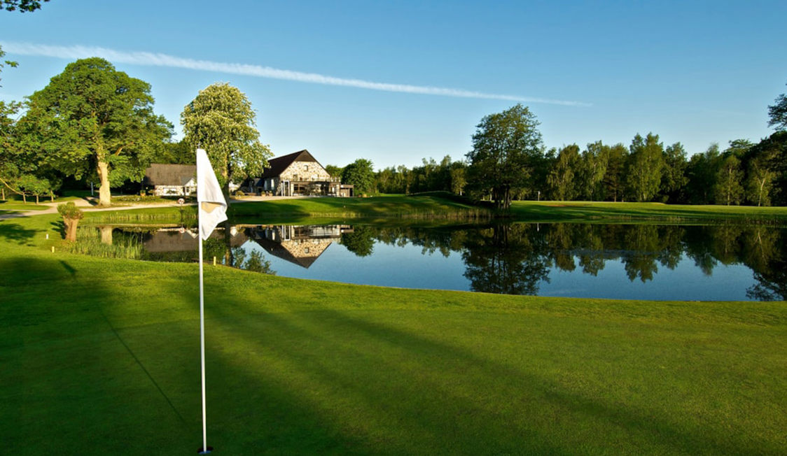 Golfbane med flag, spejlende sø og grønt græslandskab under en klar blå himmel.