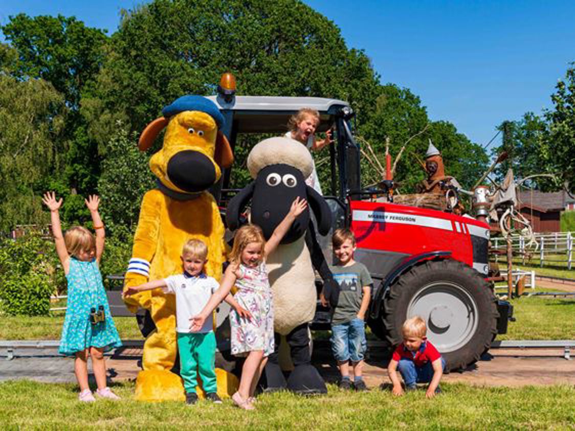 Børnegruppe poserer med store dyrekostumer ved rød traktor på en solrig dag i parken.
