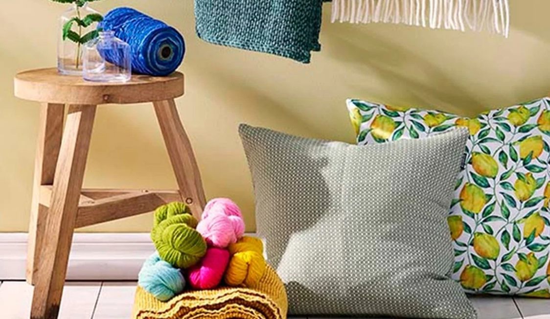 Garnnøgler i forskellige farver samt puder med citronprint på trægulv ved siden af træskammel og tæppe.