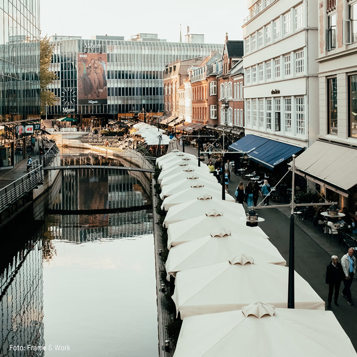 Gågade ved kanalen i Aarhus med hvide parasoller og livlige caféer på begge sider af vandet.