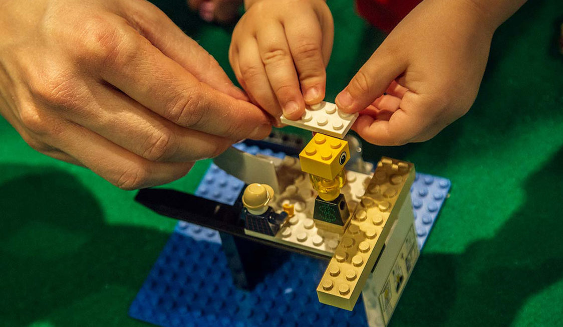 Två händer bygger en LEGO-modell på Experimentarium med små LEGO-figurer och byggklossar på en grön yta.