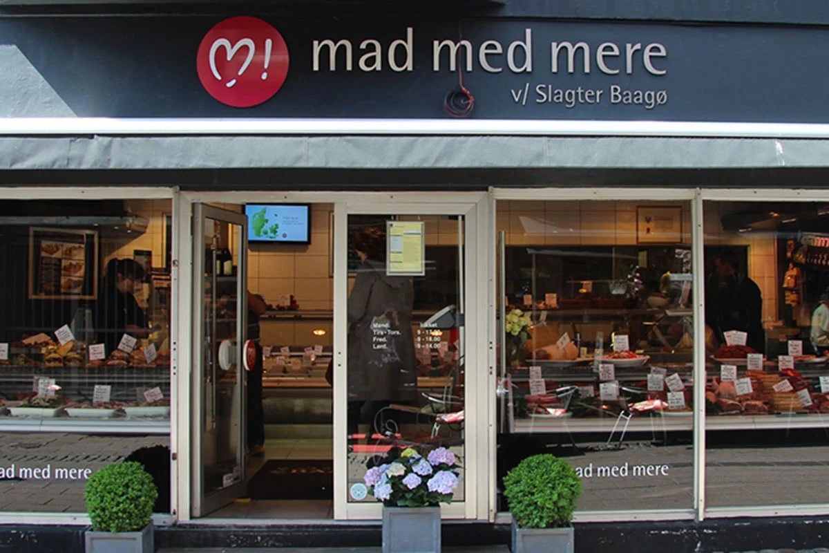Slagteri Helsingörs affärsfasad med köttprodukter i skyltfönstret och Blomsterdekorationer framför ingången.