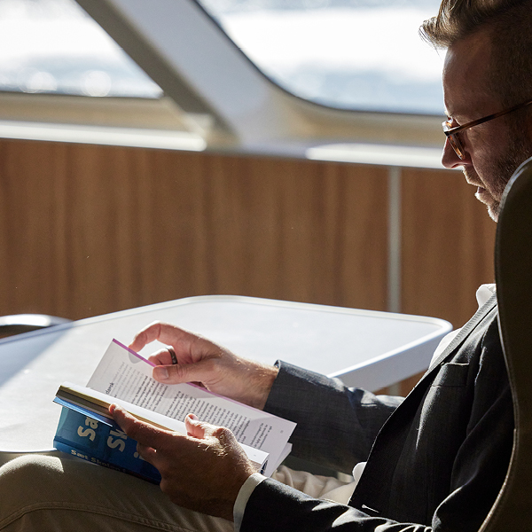 Mand i jakkesæt læser en bog ved et bord ved siden af et stort vindue ombord på en færge.