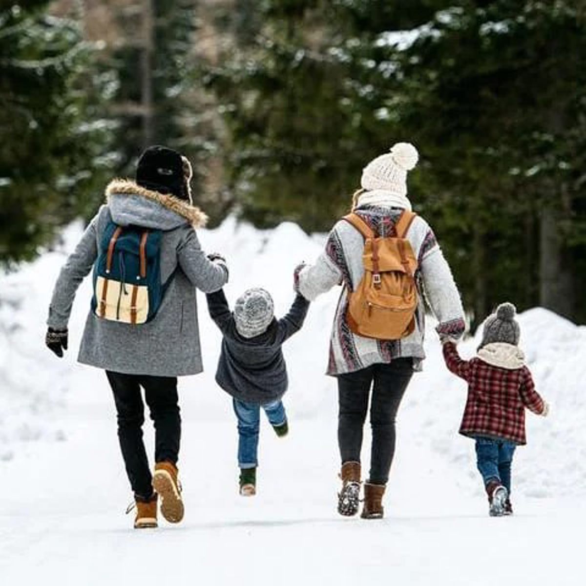 Familie går hånd i hånd på en sneklædt sti omgivet af sne og grantræer iført vintertøj og rygsække.