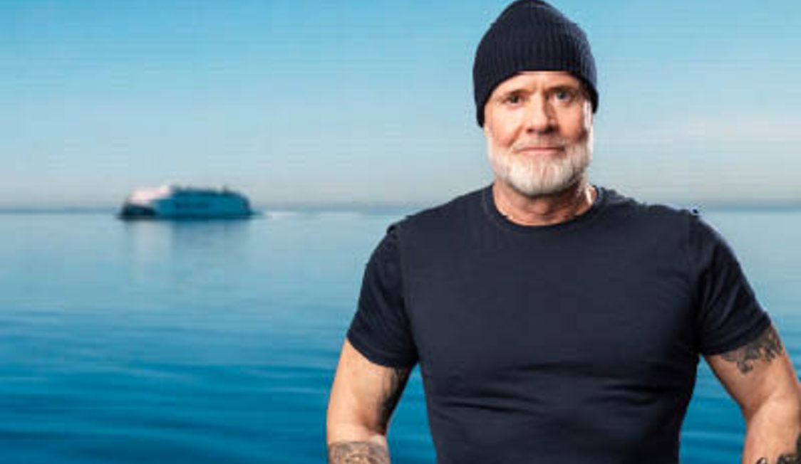 Rathsak sort t-shirt og hue står foran havet med en færge i baggrunden under klar blå himmel.