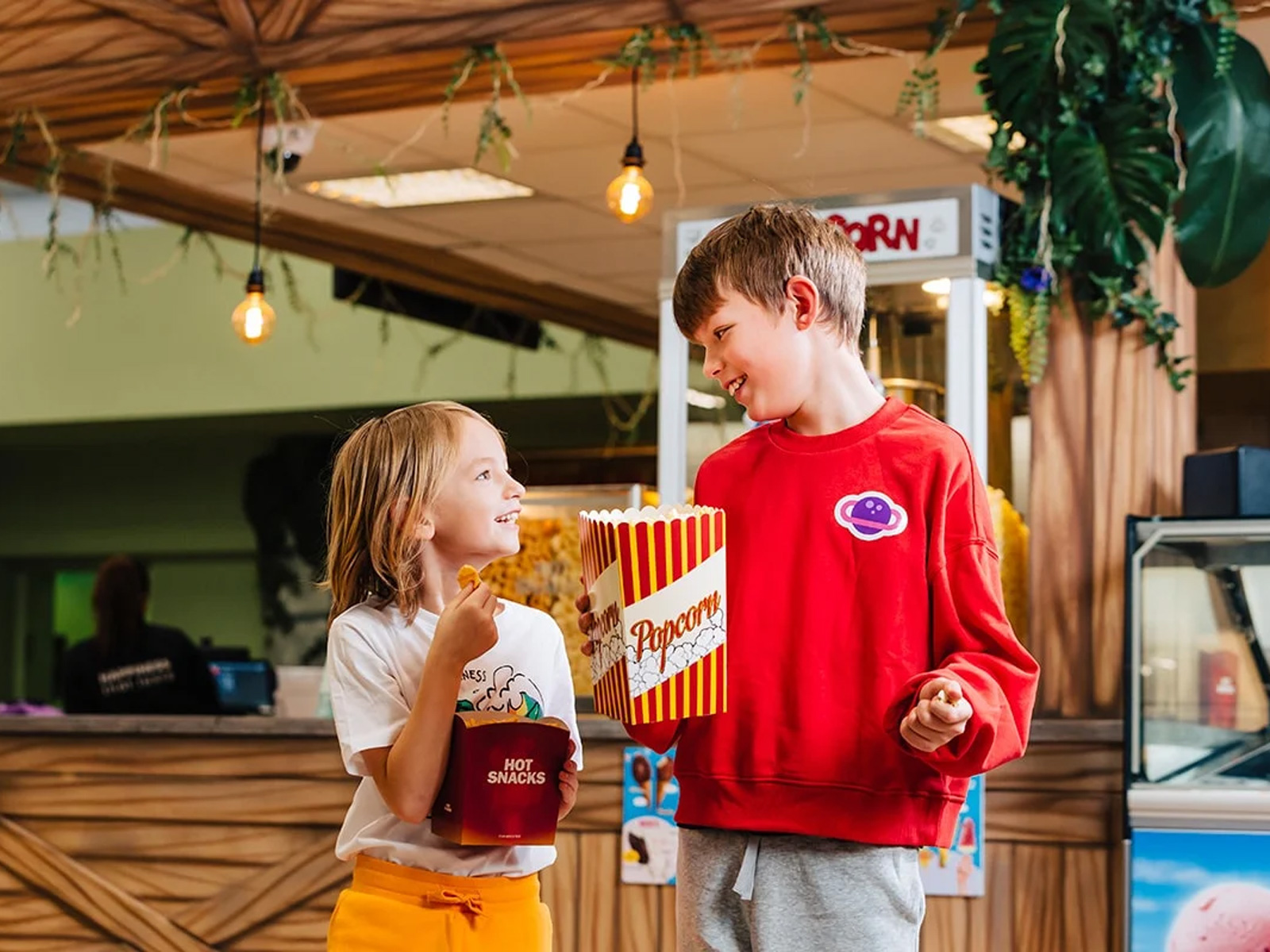 To børn deler popcorn ved biografen og smiler til hinanden under lyse lamper.