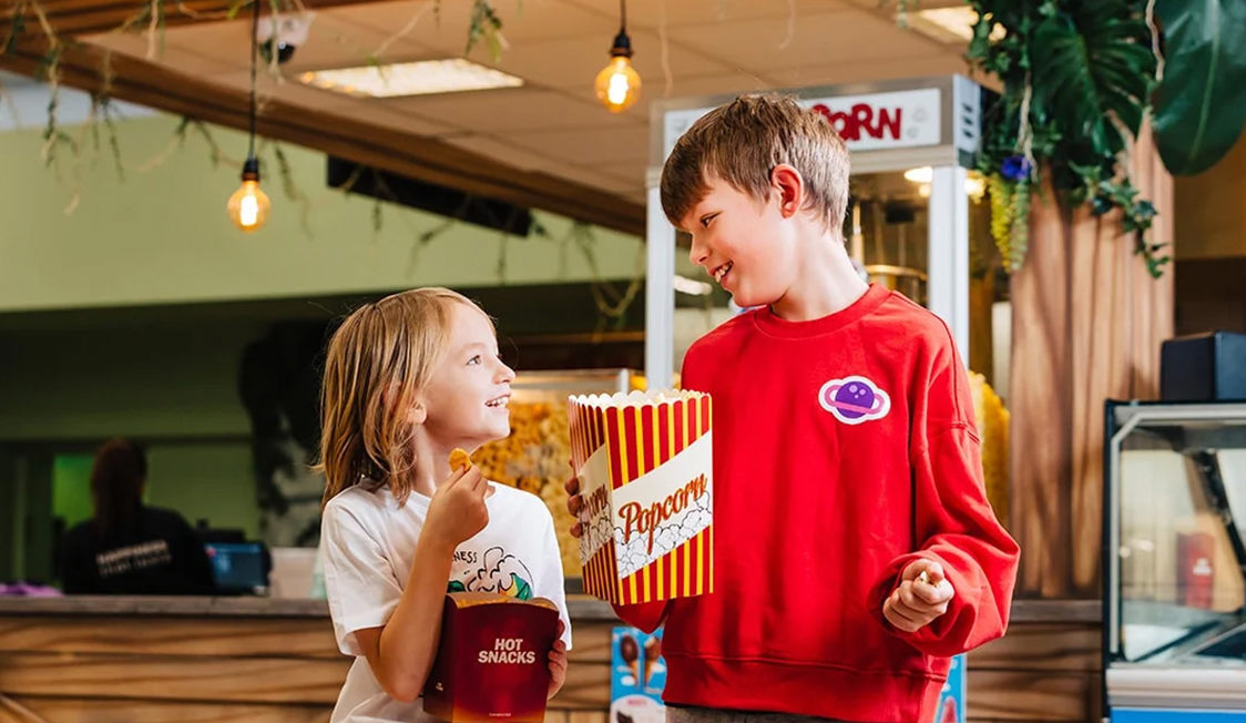 To børn deler popcorn ved biografen og smiler til hinanden under lyse lamper.
