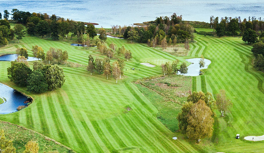 Oversigt over en frodig golfbane med klippede fairways og små søer, omkranset af træer og med udsigt til en sø i baggrunden.