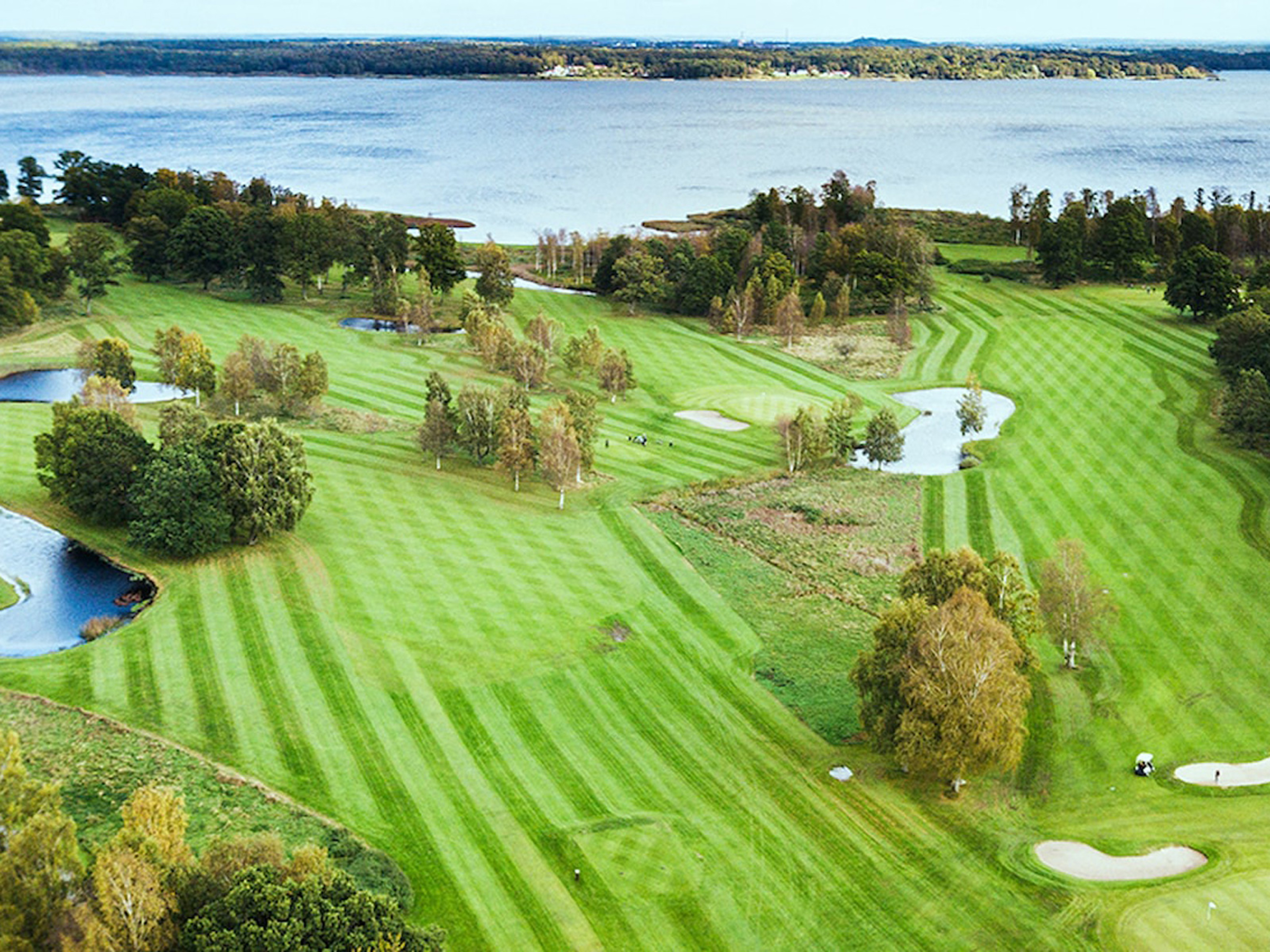 Oversigt over en frodig golfbane med klippede fairways og små søer, omkranset af træer og med udsigt til en sø i baggrunden.