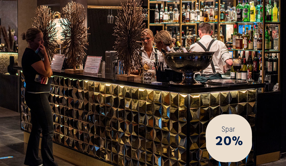 Bar med gylden mosaikfront og bartendere serverer drikkevarer, mens en person taler i telefon ved disken.