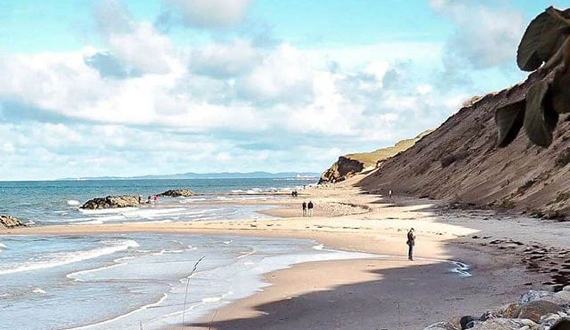 Nordsjællands Vejby strand med vågor som rullar in mot sandstranden och en klippig bakgrund under blå himmel.