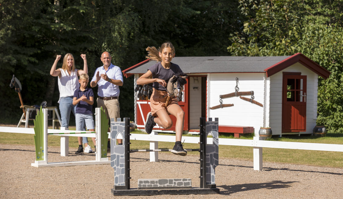 En tjej hoppar över ett hinder på en leksakshäst medan en familj hejar på i bakgrunden.
