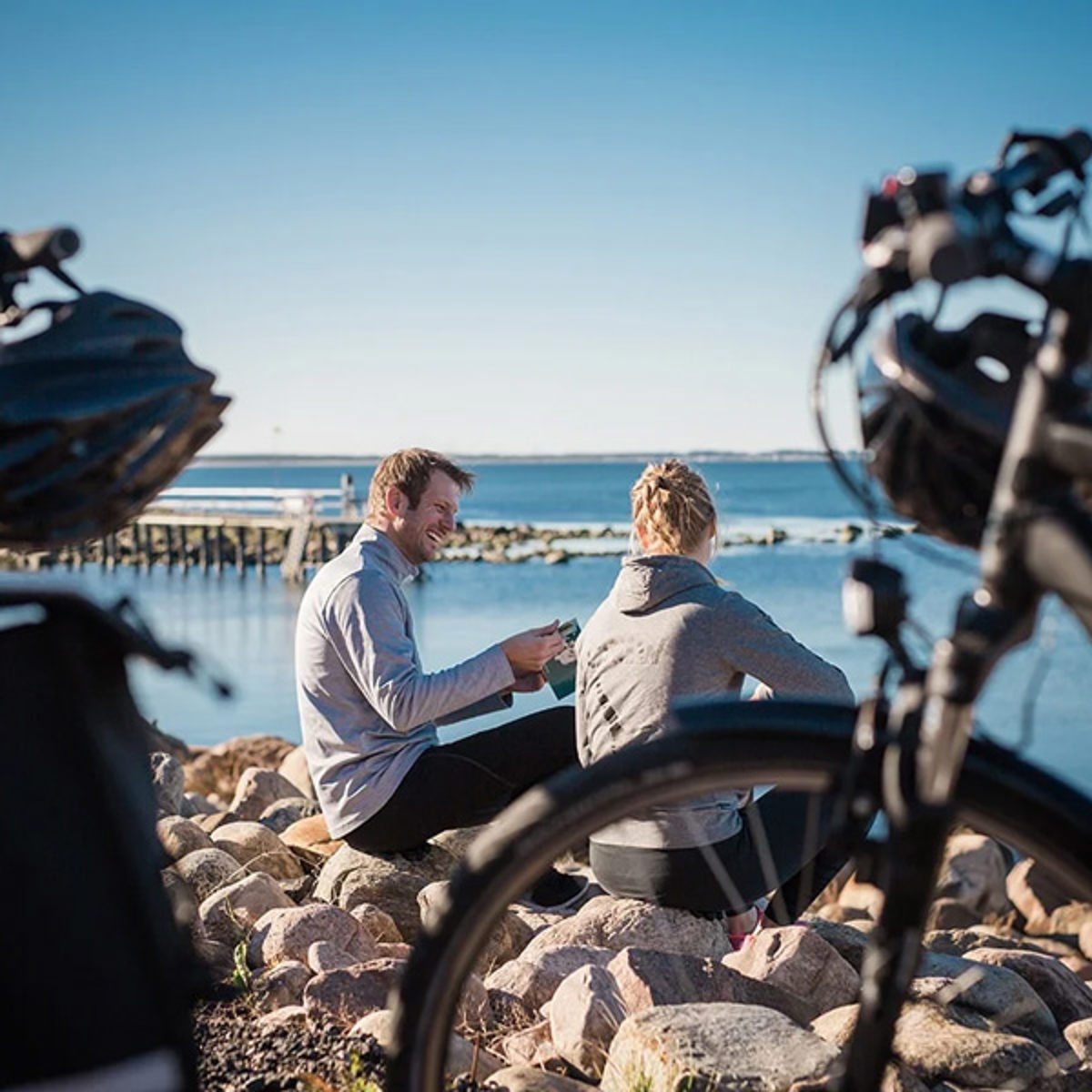To mennesker sidder på sten og hygger ved vandet med cykler i forgrunden på en solrig dag.