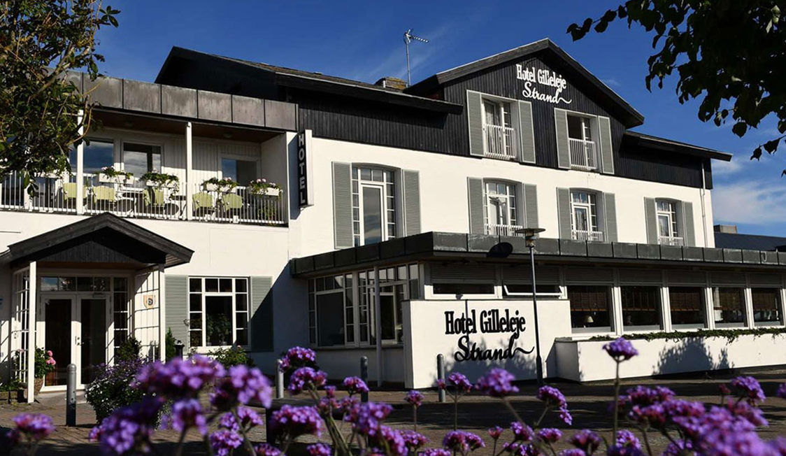 Framsidan av Hotel Gilleleje Strand med solbelysta vita väggar och balkonger med gröna växter.