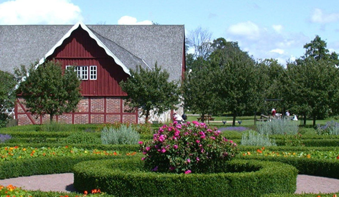 Rød bindingsværkslade med stråtag omgivet af symmetrisk anlagt have med blomsterbede og buske.