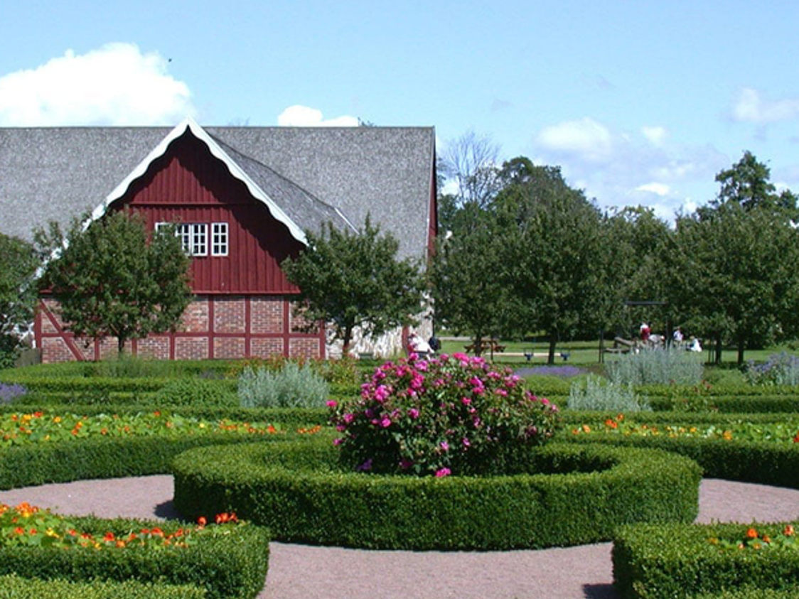 Rød bindingsværkslade med stråtag omgivet af symmetrisk anlagt have med blomsterbede og buske.