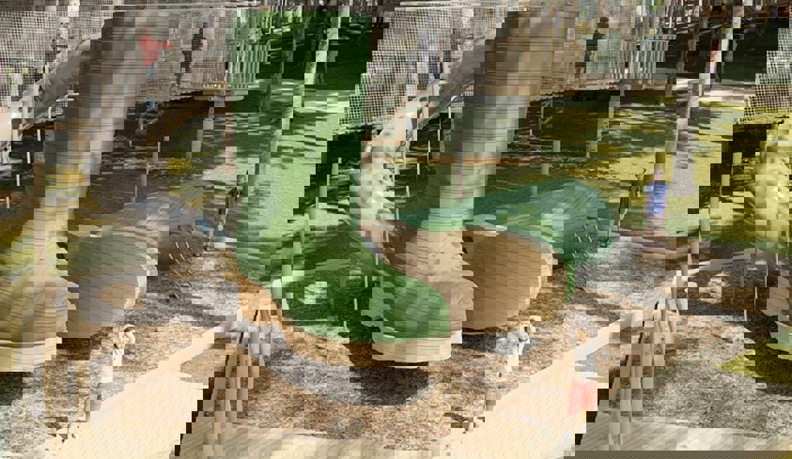 Legeplads, der ligner et par grønne støvler, med gynger og klatreudstyr i solskinsvejr.