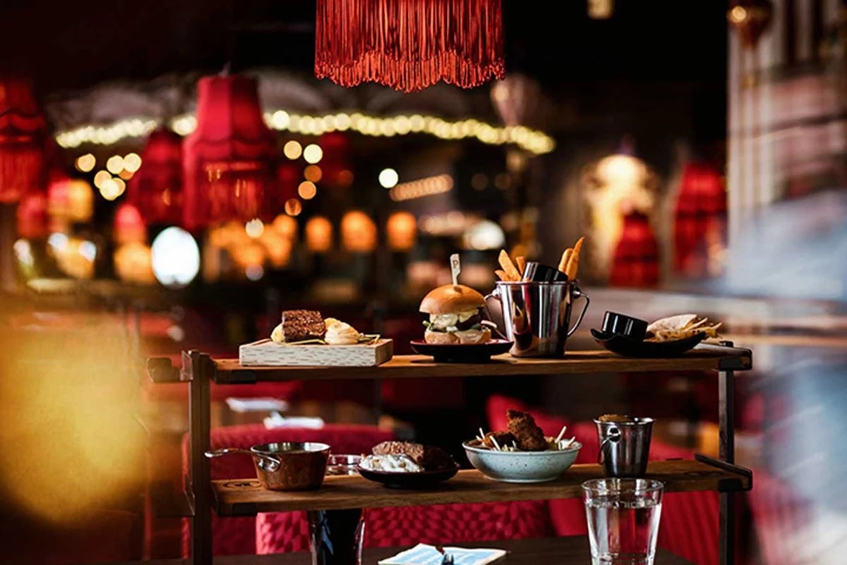 Serveringsvogn med gourmetretter og snacks i en stemningsfuld restaurant oplyst af røde lamper.