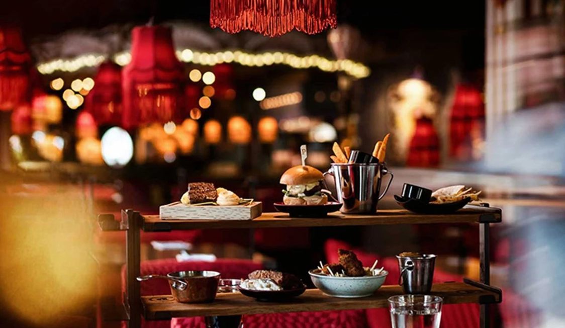 Serveringsvogn med gourmetretter og snacks i en stemningsfuld restaurant oplyst af røde lamper.