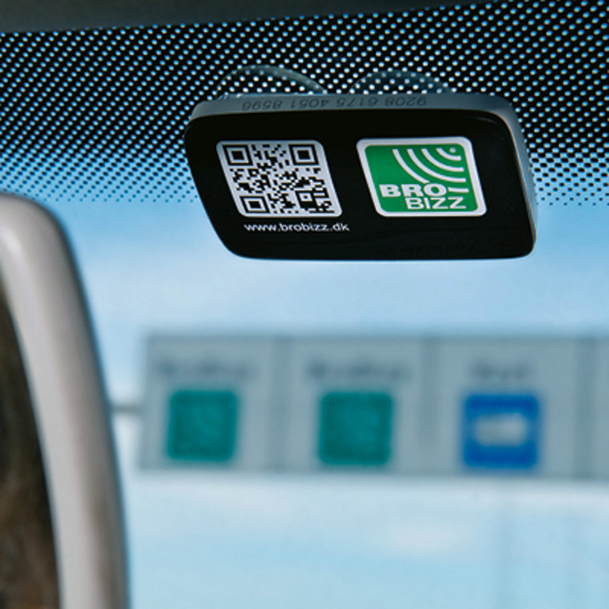 BroBizz enhed monteret på forruden af en bil, med vejskilte synlige i baggrunden, klar til elektronisk betaling.