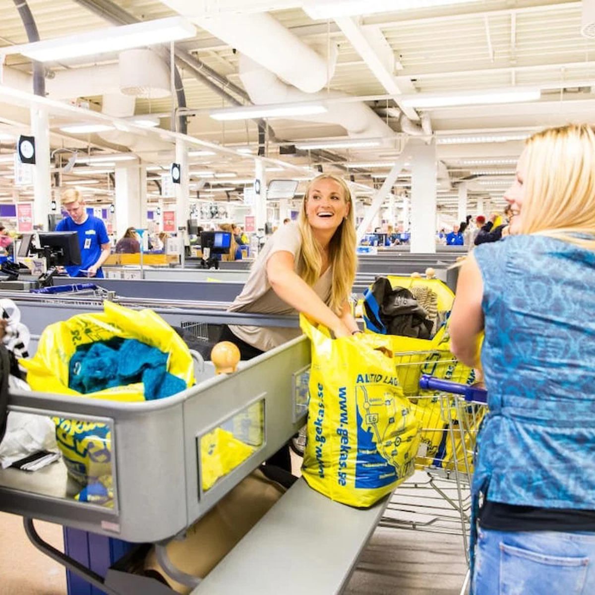 Kvinde pakker indkøb i gul indkøbspose i supermarked, mens hun taler med smilende kunder ved kassen.